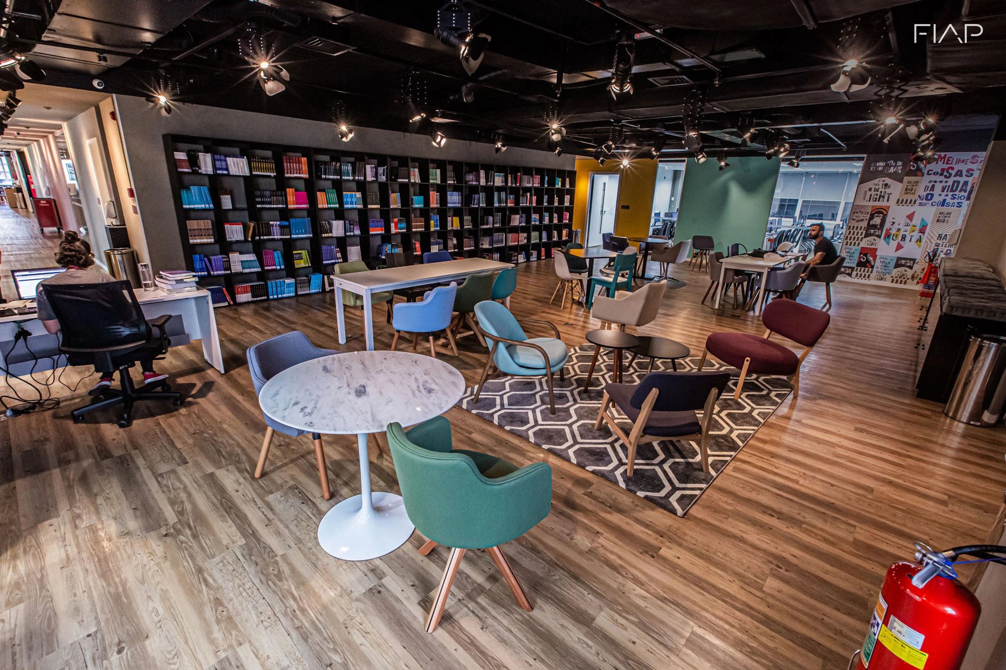 Interior da biblioteca do campus Paulista da FIAP, com mesas e cadeiras para estudo e estantes cheias de livros.