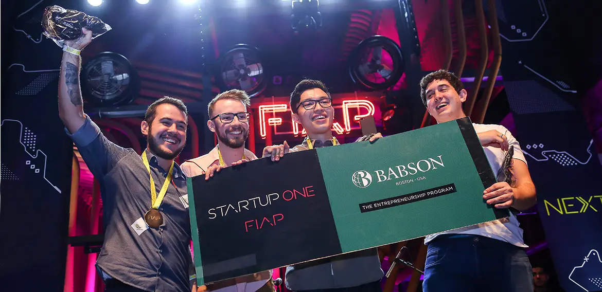 Quatro jovens empreendedores comemorando a vitória em um evento da FIAP, segurando um cheque gigante de premiação, com o logotipo STARTUP ONE FIAP e BABSON - The Entrepreneurship Program.